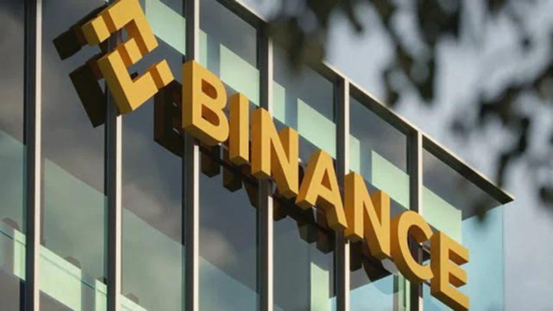 Papan nama Binance berwarna emas dipasang pada fasad kaca gedung kantor dengan latar langit biru