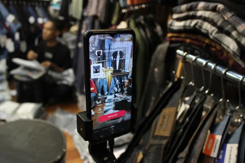Layar smartphone menampilkan persiapan siaran langsung TikTok Shop di depan gantungan pakaian kios pasar